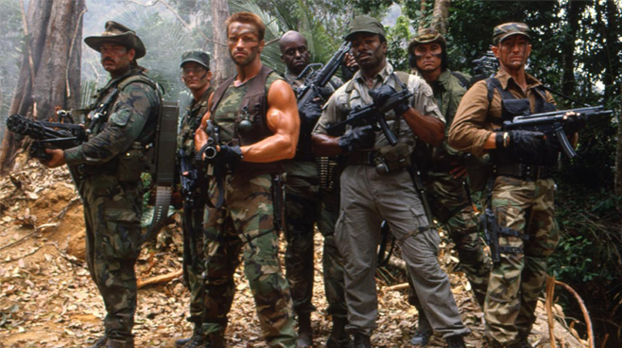 Équipe de soldats en tenue de combat dans une jungle dense, avec Arnold Schwarzenegger au centre, tenant une arme et entouré de ses coéquipiers armés.