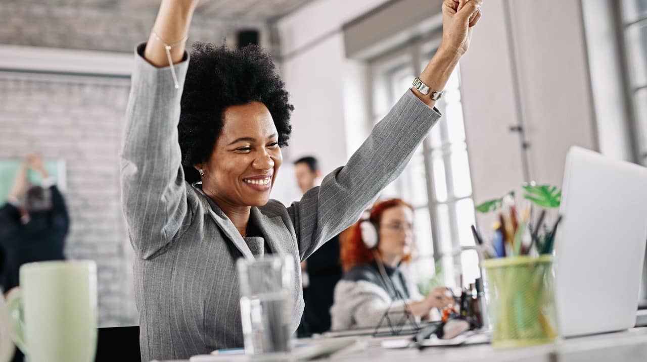 Une femme souriante en costume gris lève les bras en signe de victoire dans un environnement de bureau lumineux, célébrant un succès.