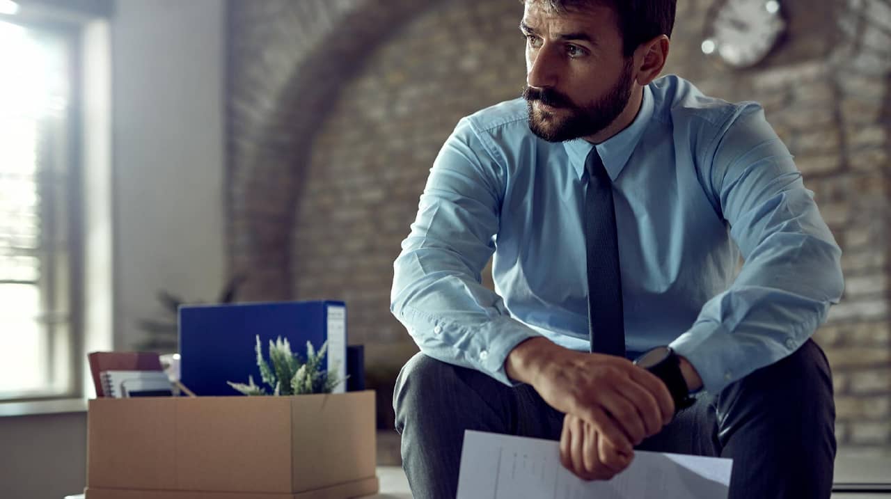 Un homme en costume, l'air pensif, assis avec un carton de bureau rempli d'effets personnels, symbolisant un licenciement.