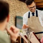 Un serveur souriant en tablier discute avec une cliente dans un restaurant chaleureux, tandis qu’un autre client est flou au premier plan.