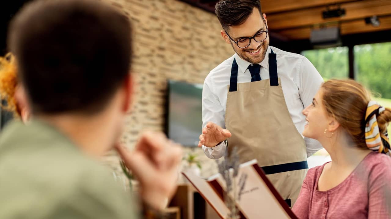 Un serveur souriant en tablier discute avec une cliente dans un restaurant chaleureux, tandis qu’un autre client est flou au premier plan.