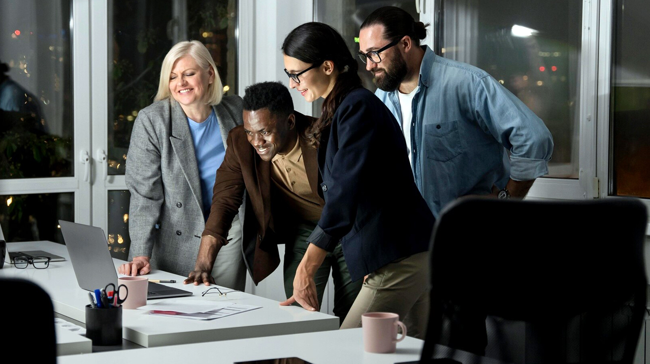 Un groupe de collègues souriants, réunis autour d’un ordinateur dans un bureau moderne. Ils semblent engagés et enthousiastes, illustrant un environnement de travail collaboratif et positif.