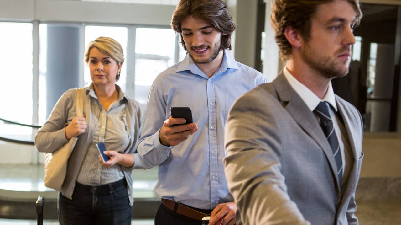 Trois professionnels en file d’attente dans un cadre formel. L’un d’eux, souriant et concentré sur son téléphone, semble sûr de lui, tandis qu’une femme en arrière-plan a une expression plus hésitante.