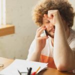 Un jeune entrepreneur assis à son bureau, visiblement stressé, la main sur le front et en pleine conversation téléphonique, symbolisant les difficultés et les doutes liés à l’échec entrepreneurial.