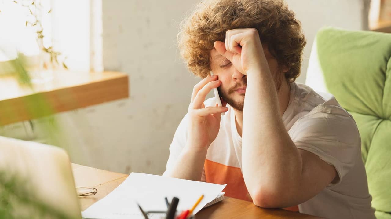Un jeune entrepreneur assis à son bureau, visiblement stressé, la main sur le front et en pleine conversation téléphonique, symbolisant les difficultés et les doutes liés à l’échec entrepreneurial.