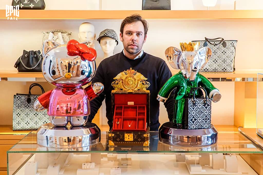 Vincent Faudemer pose entre deux statues chromées — une Hello Kitty rose et un Babolex vert — exposées dans une boutique de luxe, devant des sacs griffés.